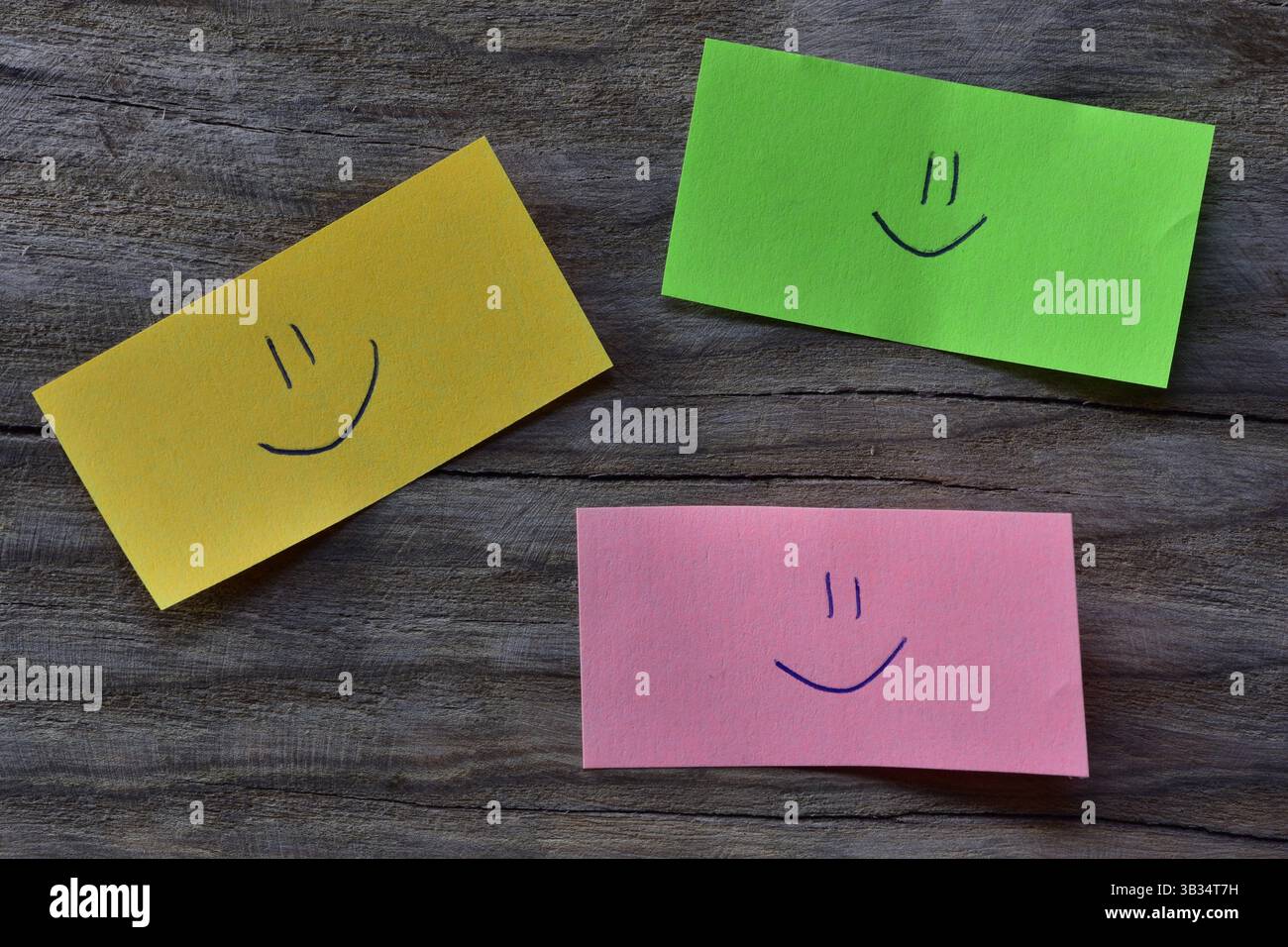 Visage souriant sur note collante verte, jaune et rose sur fond texturé de planche de bois. Un stylo dessine un visage souriant sur du papier à notes. Banque D'Images