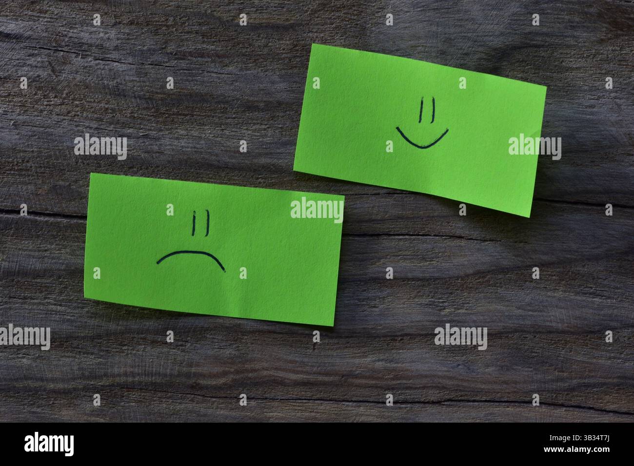 Visage souriant et visage triste sur note collante verte sur fond texturé de planche de bois. Un stylo dessine un visage souriant et un visage triste sur du papier à lettres. Banque D'Images
