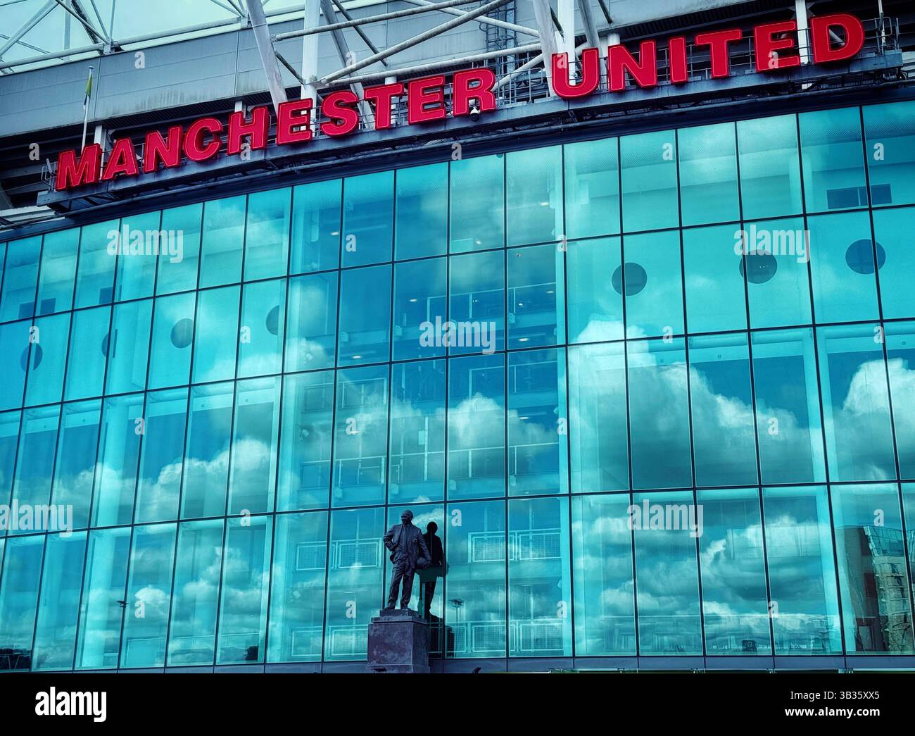 Statue de Sir Matt Busby au stade de football Old Trafford. Statue et reflet avec des nuages et le ciel bleu reflété dans les fenêtres de l'avant du stade. Manthorax - Image de stock capturée avec un smartphone