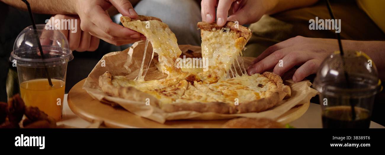 Gros plan des mains tirant des tranches de pizza au fromage d'un plateau en bois, avec des verres de jus et des hamburgers en arrière-plan. Fromage s'étirant magnifiquement des tranches de pizza Banque D'Images