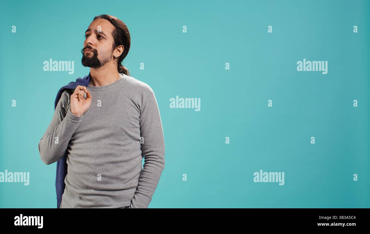 Homme tenant la veste dans une main tout en regardant autour, contemplant, créant un sentiment de curiosité. Personne tenant le manteau montrant la curiosité pour l'environnement, fond de studio, caméra B. Banque D'Images