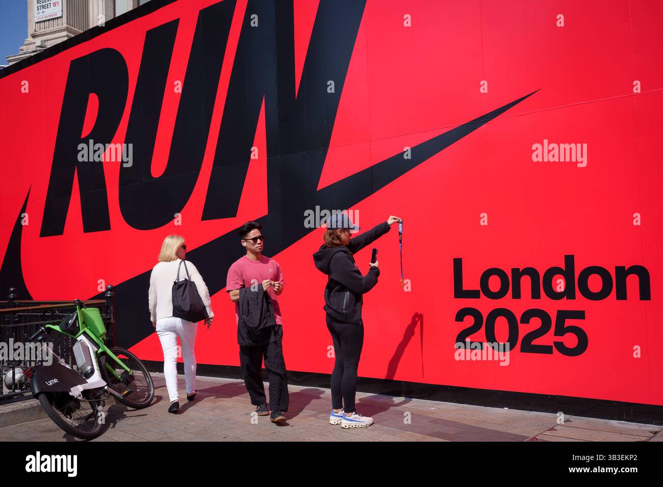 Un jour après le marathon de Londres 2025, les clients passent devant une finisseuse de course qui photographie sa propre médaille sous un panneau d'affichage à thème devant Nike Town, célébrant la course à pied et l'athlétisme, le 28 avril 2025, à Londres, en Angleterre. Banque D'Images