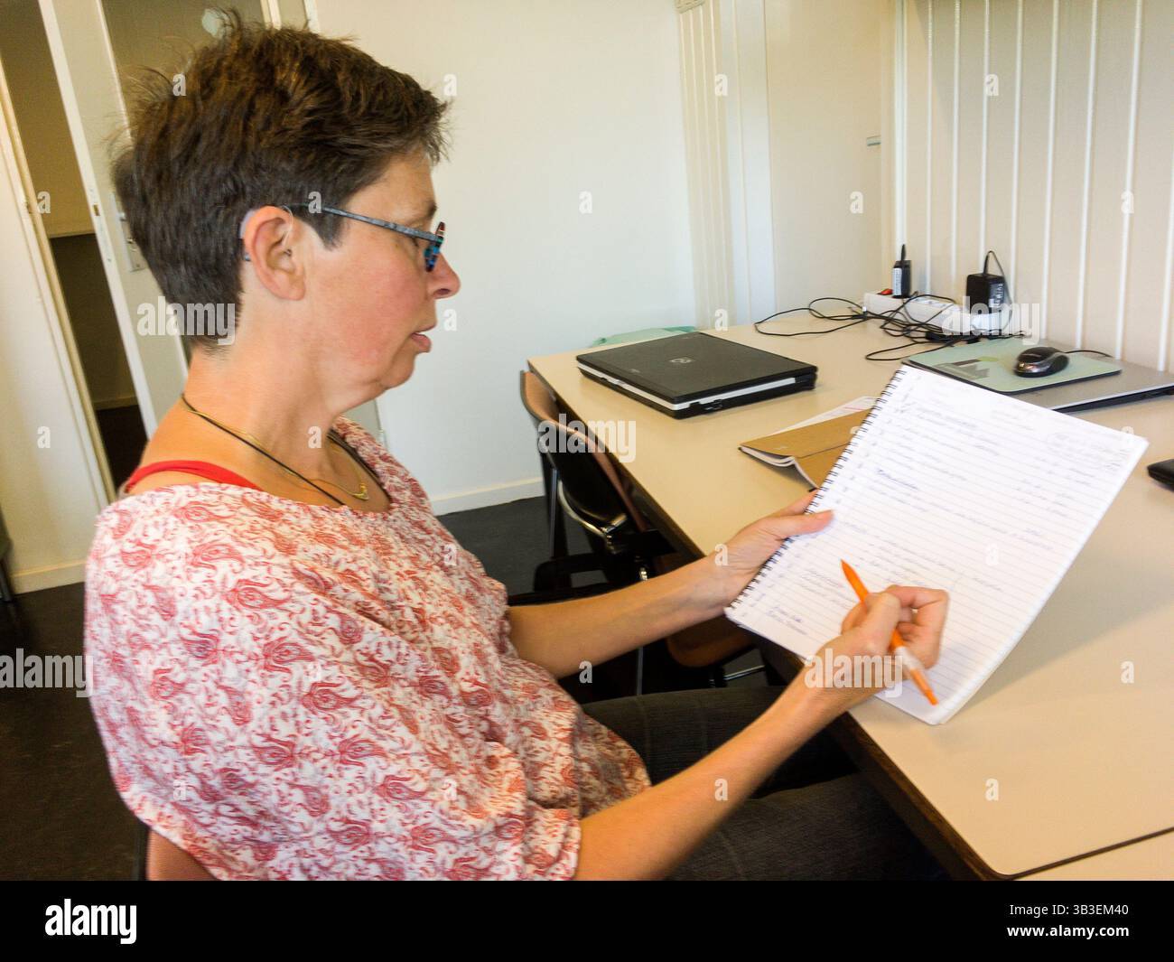 Femme adulte mature portant des lunettes travaillant derrière son bureau pour traiter des notes sur un cahier écrit manuellement. Tilburg, pays-Bas. MR=OUI Banque D'Images