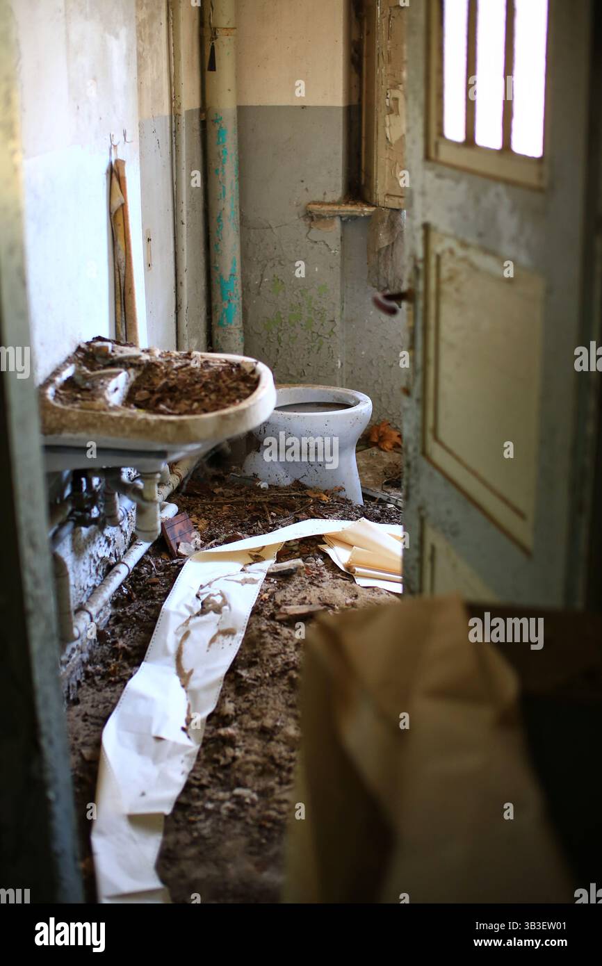 Salle de bain abandonnée avec un lavabo rempli de débris, toilettes délabrées et sol couvert de saleté avec du papier dans un espace en ruine. Banque D'Images