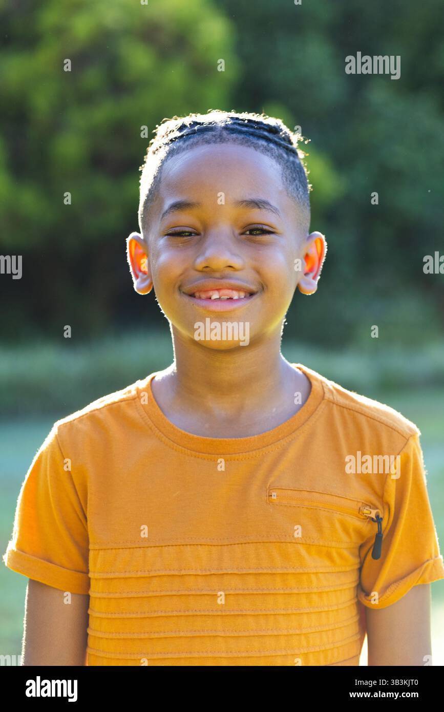 Garçon souriant debout dans le parc portant une chemise orange avec des cornrows tressés, avec un ciel ensoleillé Banque D'Images