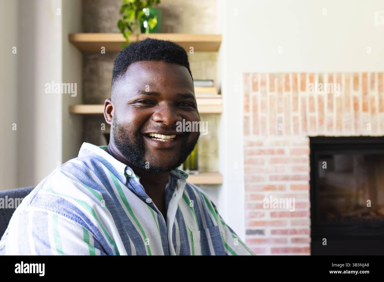 Souriant homme afro-américain mi-adulte assis dans le salon, avec cheminée en brique et étagères Banque D'Images