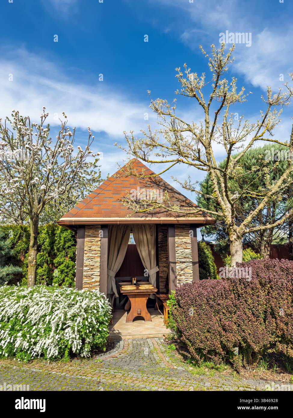 Aménagement de jardin magnifique avec un gazebo en pierre, des arbres en fleurs et des buissons au printemps. Gazebo de jardin élégant avec toit carrelé au milieu des floraisons Banque D'Images