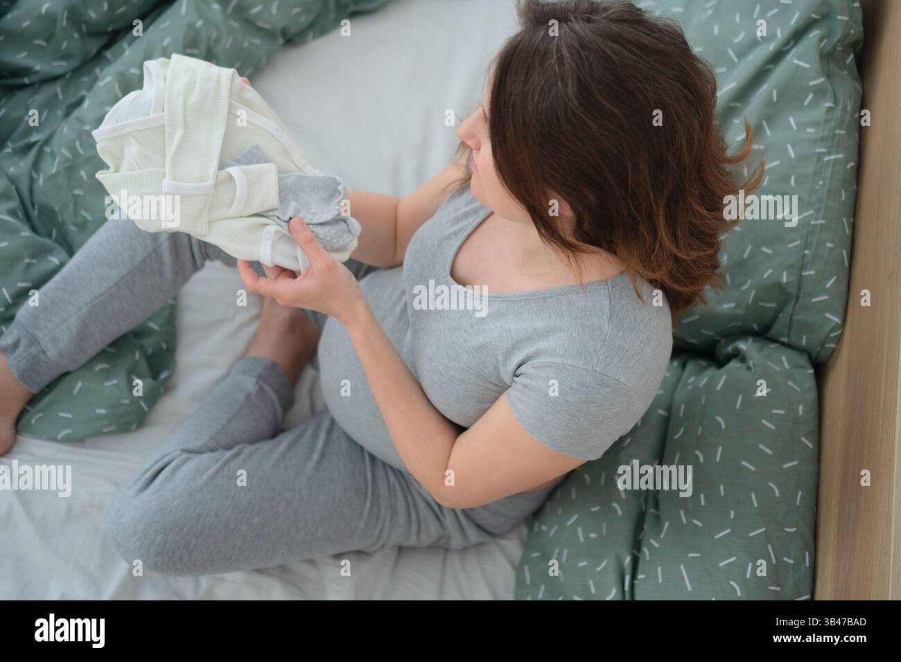 Une femme enceinte brillante est assise les jambes croisées, entourée de vêtements de bébé, touchant doucement son ventre. Un moment tranquille d’amour et d’anticipation. Banque D'Images
