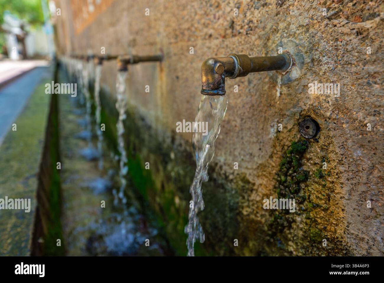 De l'eau coule d'une rangée de vieux becs métalliques installés dans un mur de fontaine en pierre, capturé en lumière douce du jour avec une faible profondeur de champ et des détails texturés Banque D'Images