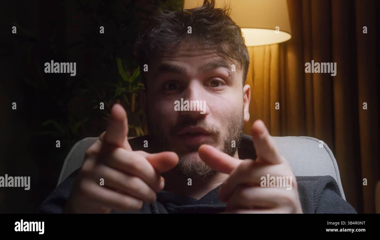 Jeune vlogger masculin parlant directement à la caméra avec des gestes expressifs de la main dans un éclairage intérieur confortable, créant du contenu attrayant pour les médias sociaux Banque D'Images