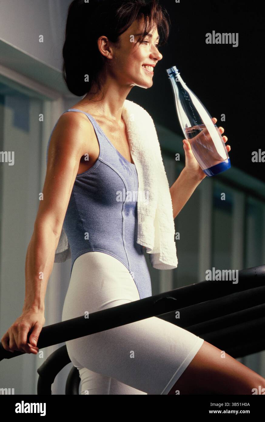 Jeune femme buvant de l'eau embouteillée pendant la formation de fitness sur le tapis roulant de gymnase. Banque D'Images