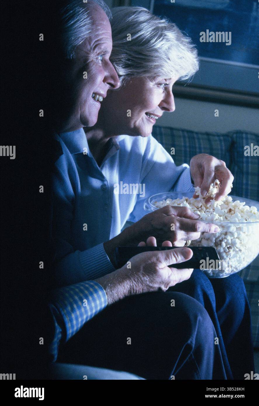 Homme senior et femme mangeant du pop-corn à l'intérieur et regardant la télévision. Banque D'Images