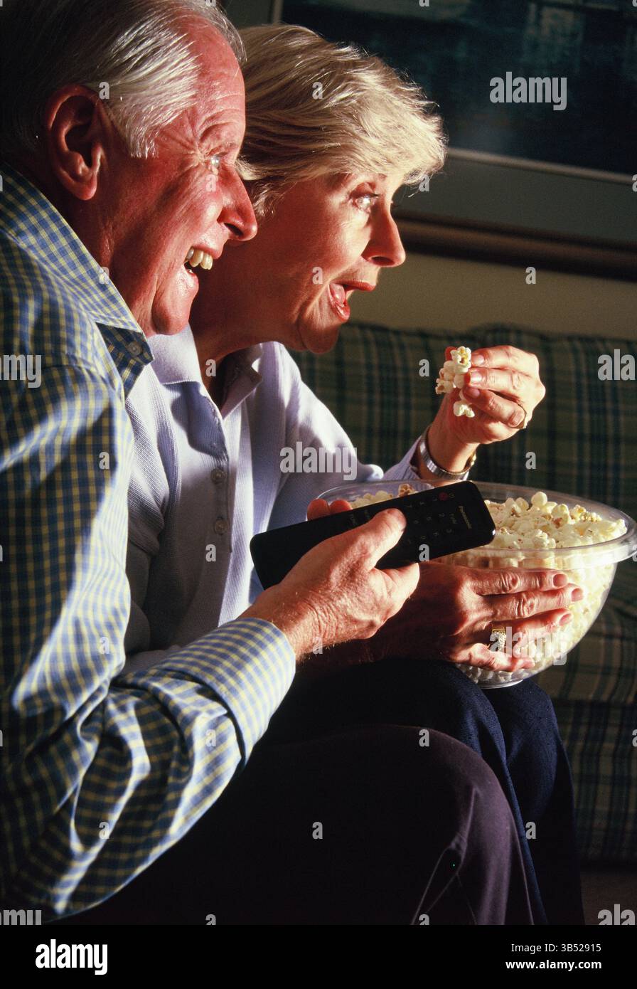 Homme senior et femme mangeant du pop-corn à l'intérieur et regardant la télévision. Banque D'Images