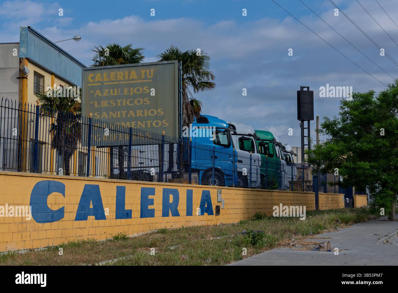 Zone de stationnement industriel avec des véhicules de construction près de la clôture à maillons de chaîne, affichant des panneaux pour les matériaux de carrelage et de plomberie Banque D'Images