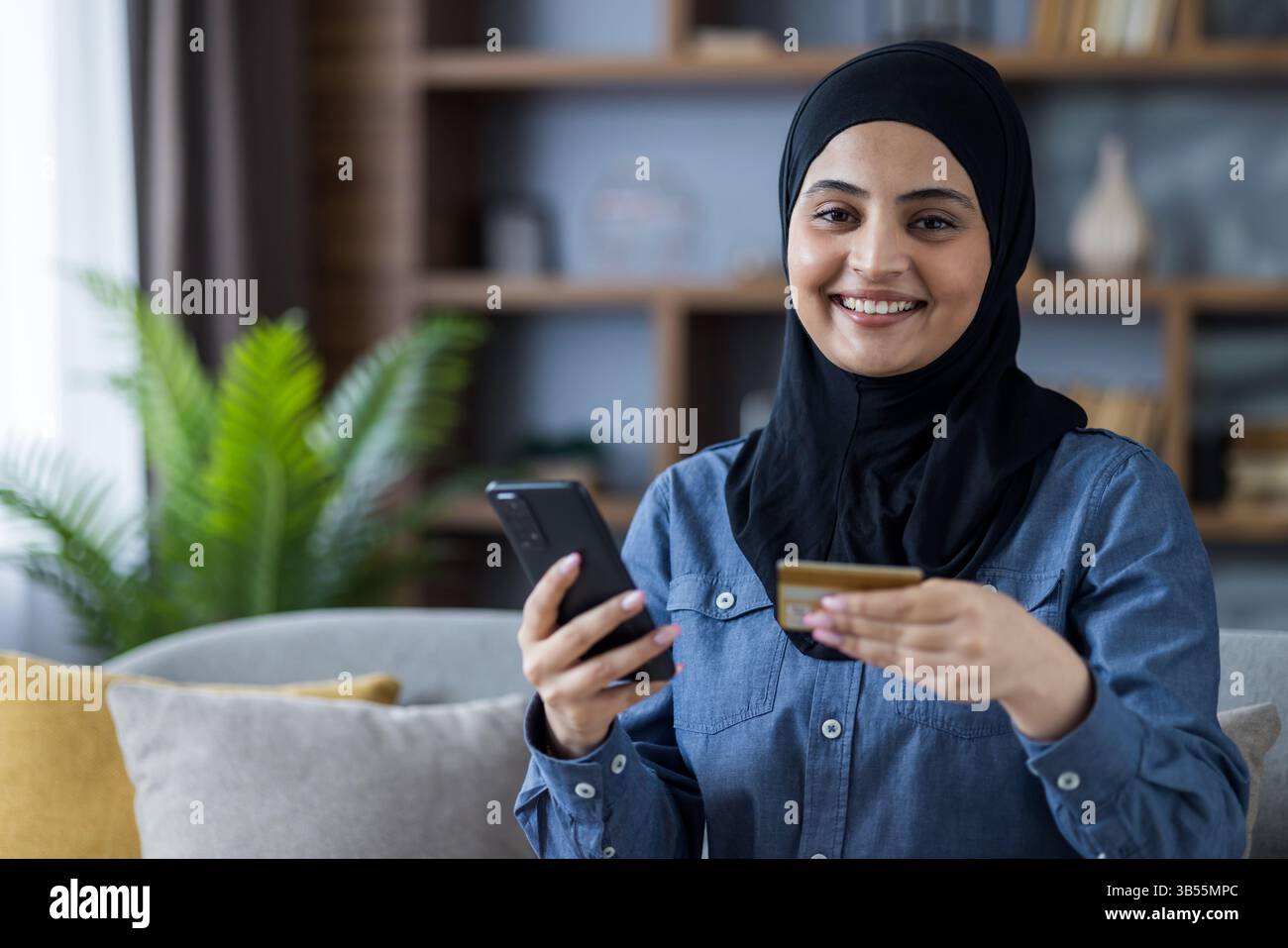 Portrait d'une jeune femme islamique dans un hijab assis sur le canapé à la maison, tenant une carte de crédit et un téléphone portable, regardant la caméra avec le sourire. Banque D'Images