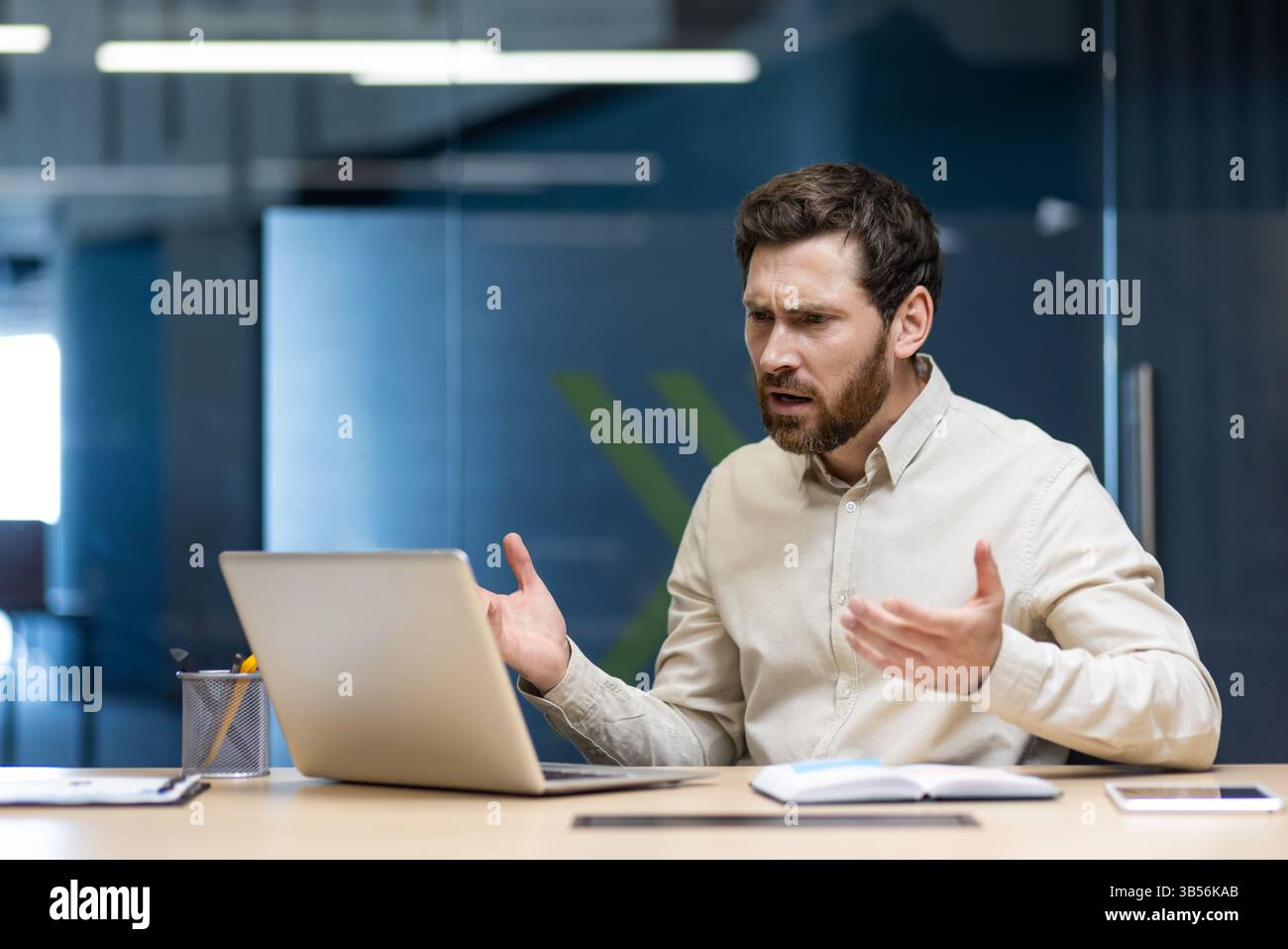 Frustré et triste jeune homme d'affaires assis au bureau dans le bureau et parlant sur l'appel vidéo sur ordinateur portable geste avec les mains. Banque D'Images