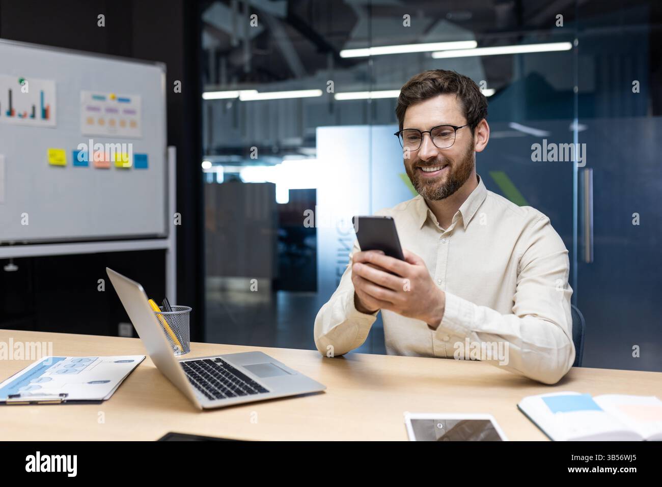 Un jeune homme souriant travaille au bureau sur un ordinateur portable, est assis derrière un bureau sur un tableau blanc avec des autocollants et utilise un téléphone portable dans ses mains. Banque D'Images