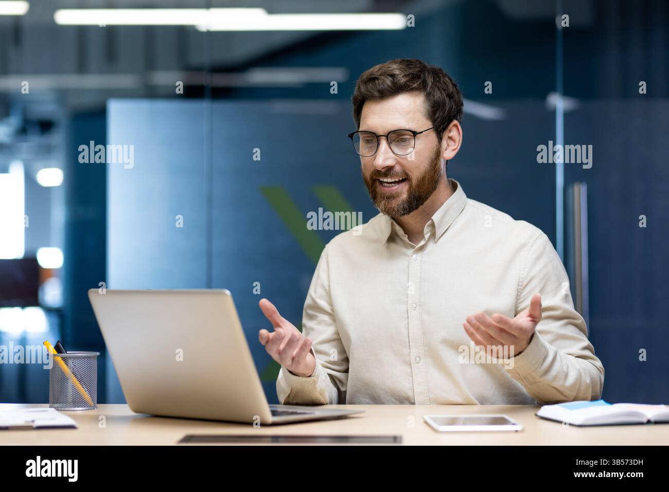 Un jeune homme d'affaires prospère est assis dans le bureau à la table de travail et dessine sur la vidéoconférence en ligne, faisant des gestes avec ses mains. Banque D'Images
