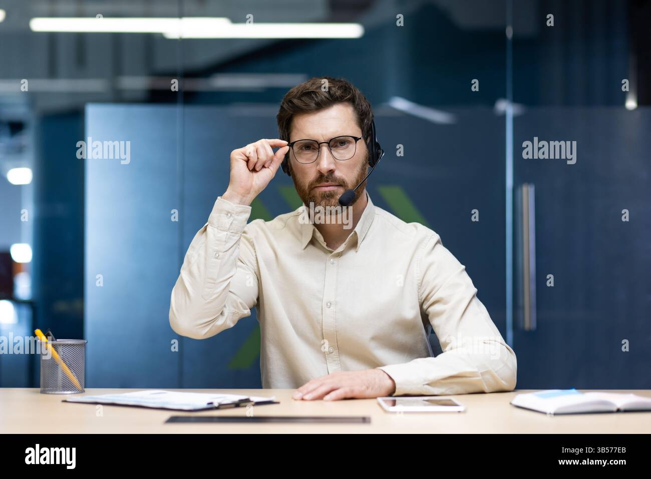 Portrait d'un jeune homme sérieux assis devant la caméra dans un casque dans le bureau, regardant la caméra et tenant des lunettes. Banque D'Images