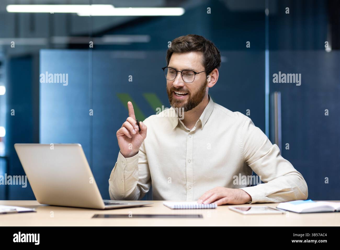 Jeune homme d'affaires prospère assis dans le bureau à la table et parlant sur un appel vidéo à travers un ordinateur portable, faisant des gestes avec ses mains et montrant un index vers le haut. Banque D'Images