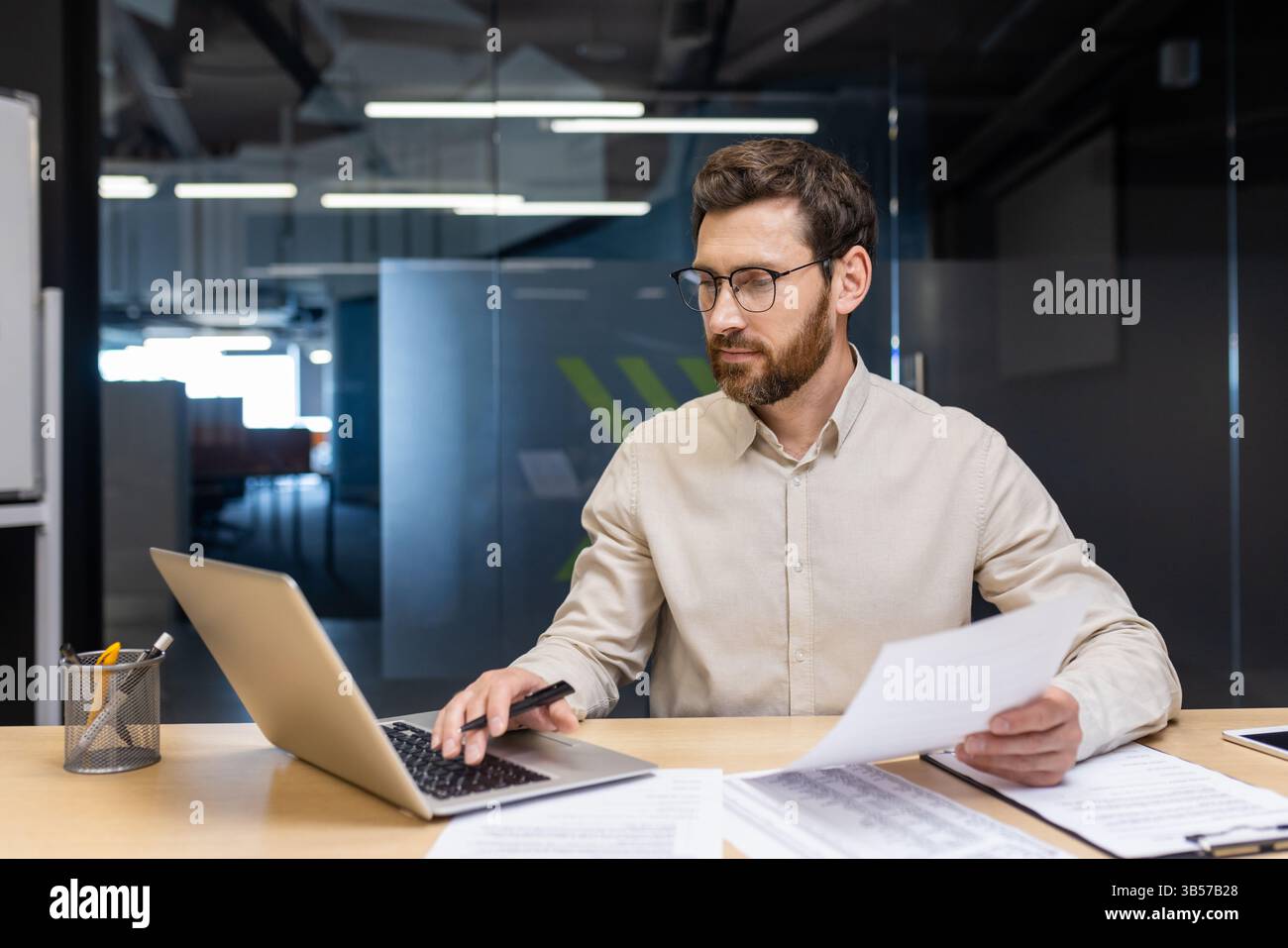 Un jeune homme concentré est assis dans le bureau à un bureau, tenant des documents dans ses mains et travaillant sur un ordinateur portable. Banque D'Images