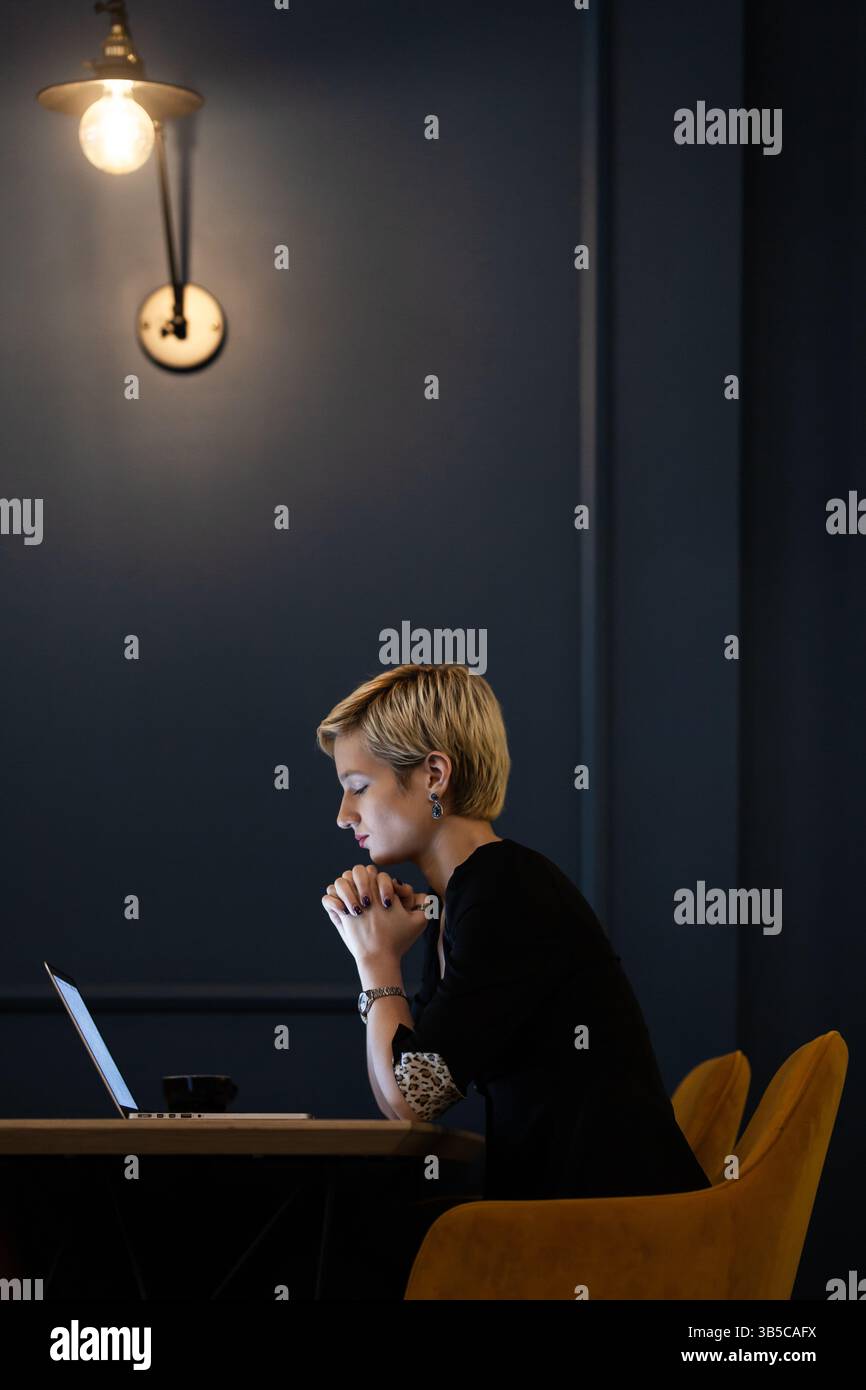 Une femme professionnelle aux cheveux blonds courts se concentre sur son ordinateur portable dans un bureau moderne, soulignant le dévouement et l'esthétisme sophistiqué du lieu de travail Banque D'Images