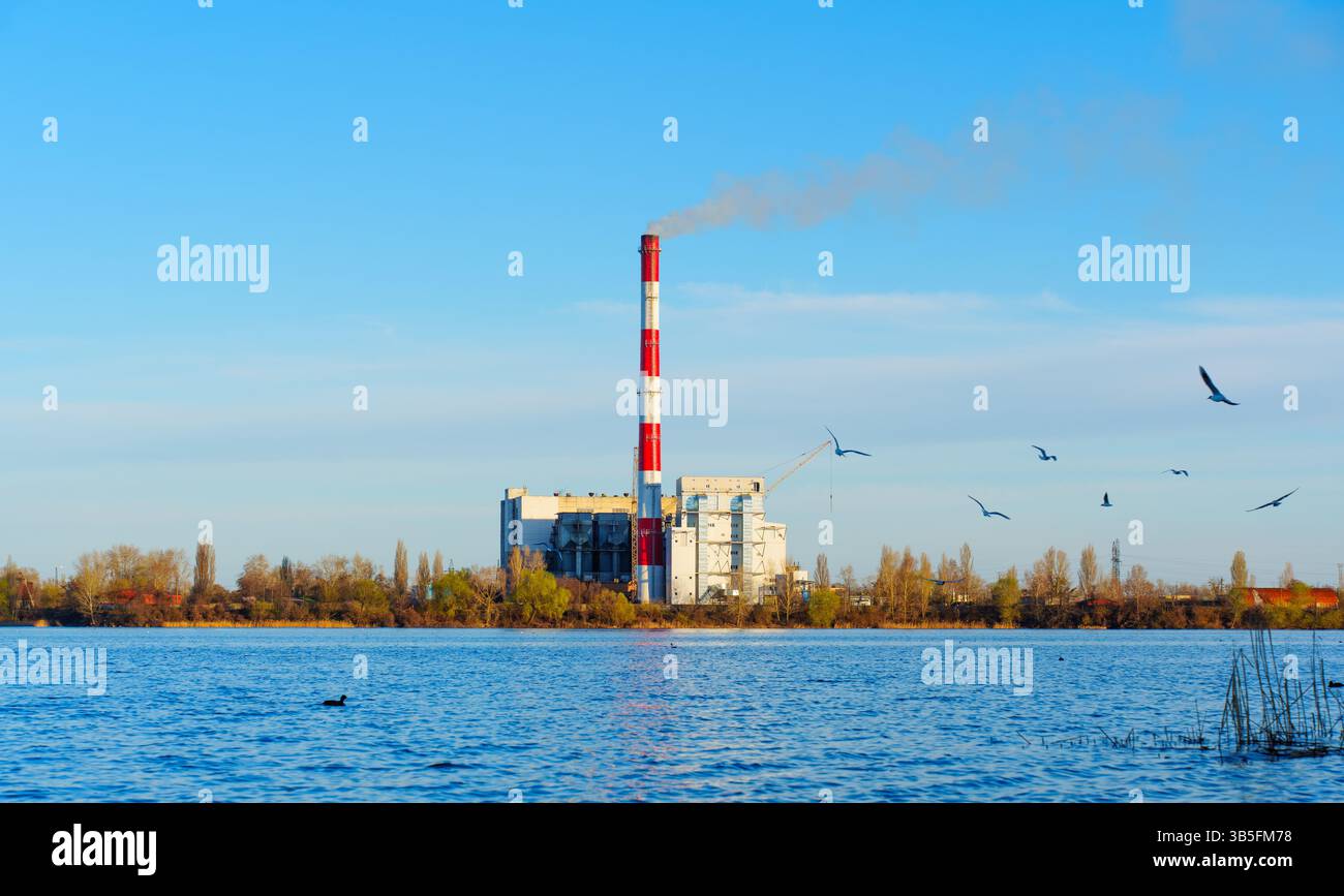L'usine d'incinération des déchets est située au bord d'un lac, avec une grande cheminée émettant de la fumée contre un ciel bleu vif, mettant en valeur le concept de gestion des déchets Banque D'Images