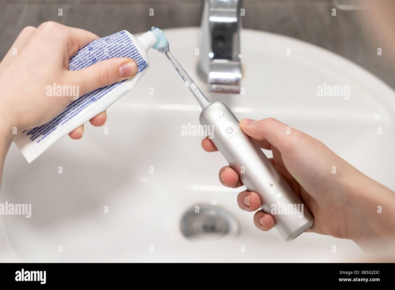 Gros plan d'une main appliquant du dentifrice sur les poils d'une brosse à dents électrique au-dessus d'un évier de salle de bain blanc, symbolisant l'auto-soin moderne et es Banque D'Images