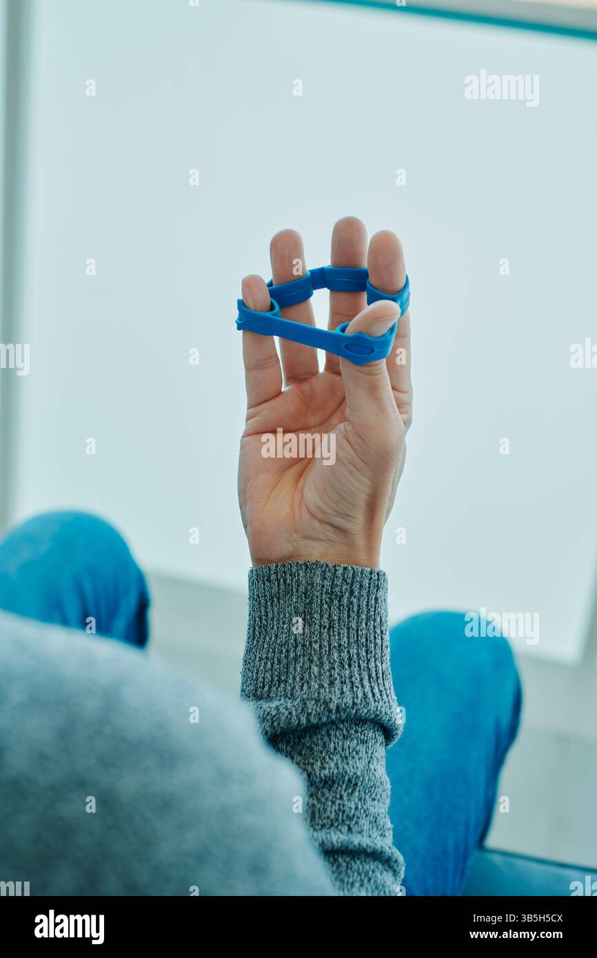 un homme utilise une bande élastique bleue pour exercer ses doigts lorsqu'il est assis sur une chaise Banque D'Images