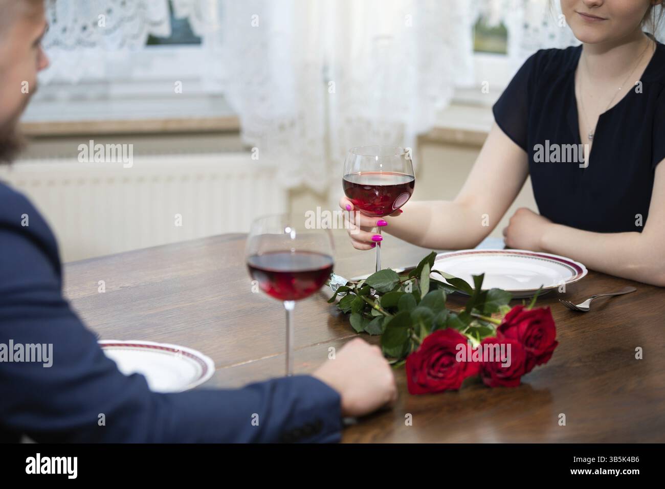 Il y a un bouquet de roses rouges sur une table en bois. À côté d'eux se trouvent des assiettes vides et deux verres de vin rouge. Vous pouvez voir des fragments des silhouettes Banque D'Images
