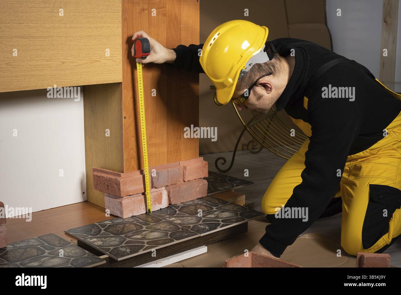 Un ouvrier de la construction en vêtements de travail, agenouillé, mesure les carreaux décoratifs rétractables en forme de vieille brique rouge avec un ruban à mesurer Banque D'Images