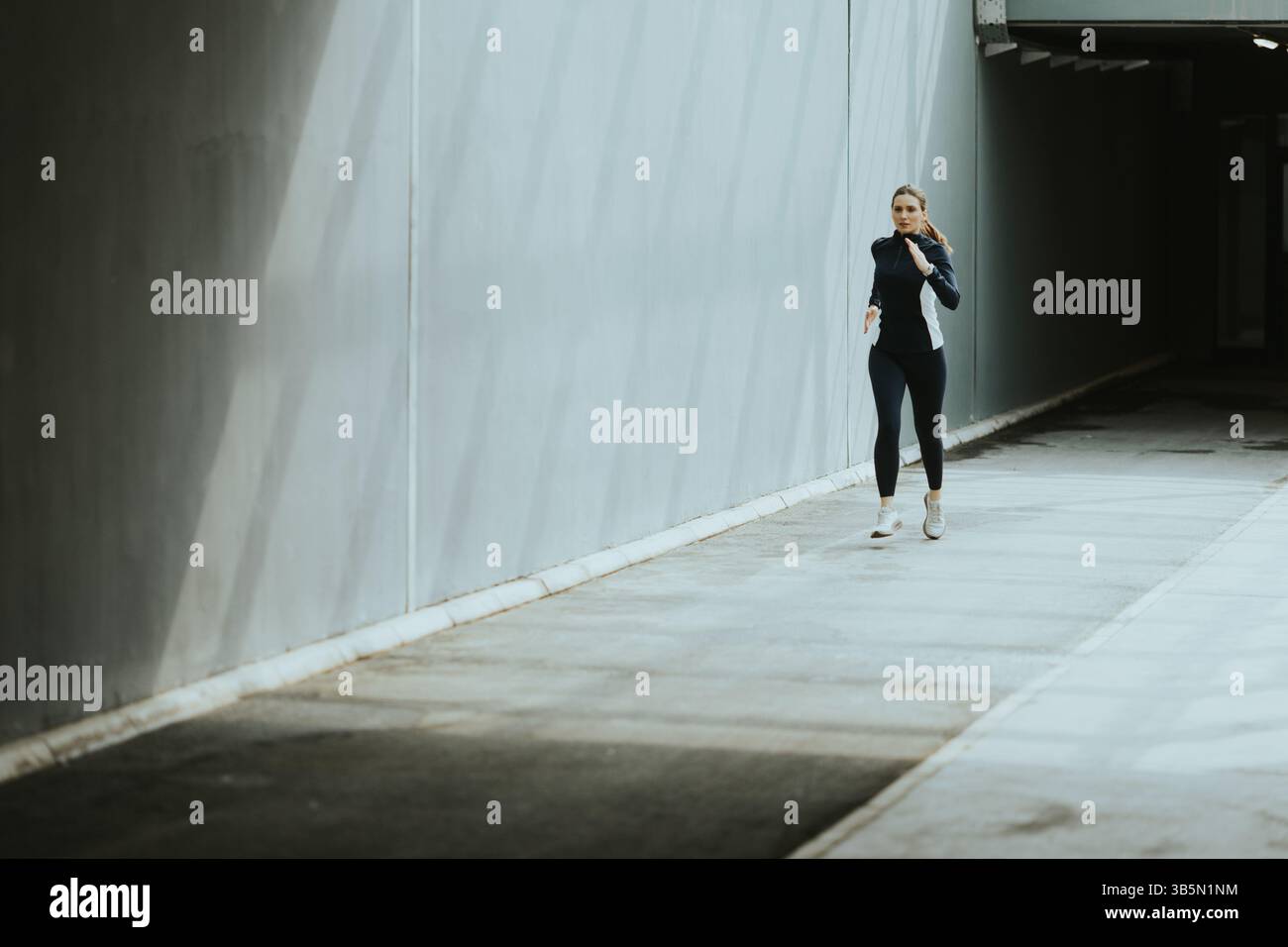 Une femme fait un jogging rapide le long d'un étroit couloir ensoleillé, profitant d'une course matinale revigorante dans un environnement urbain paisible. Banque D'Images