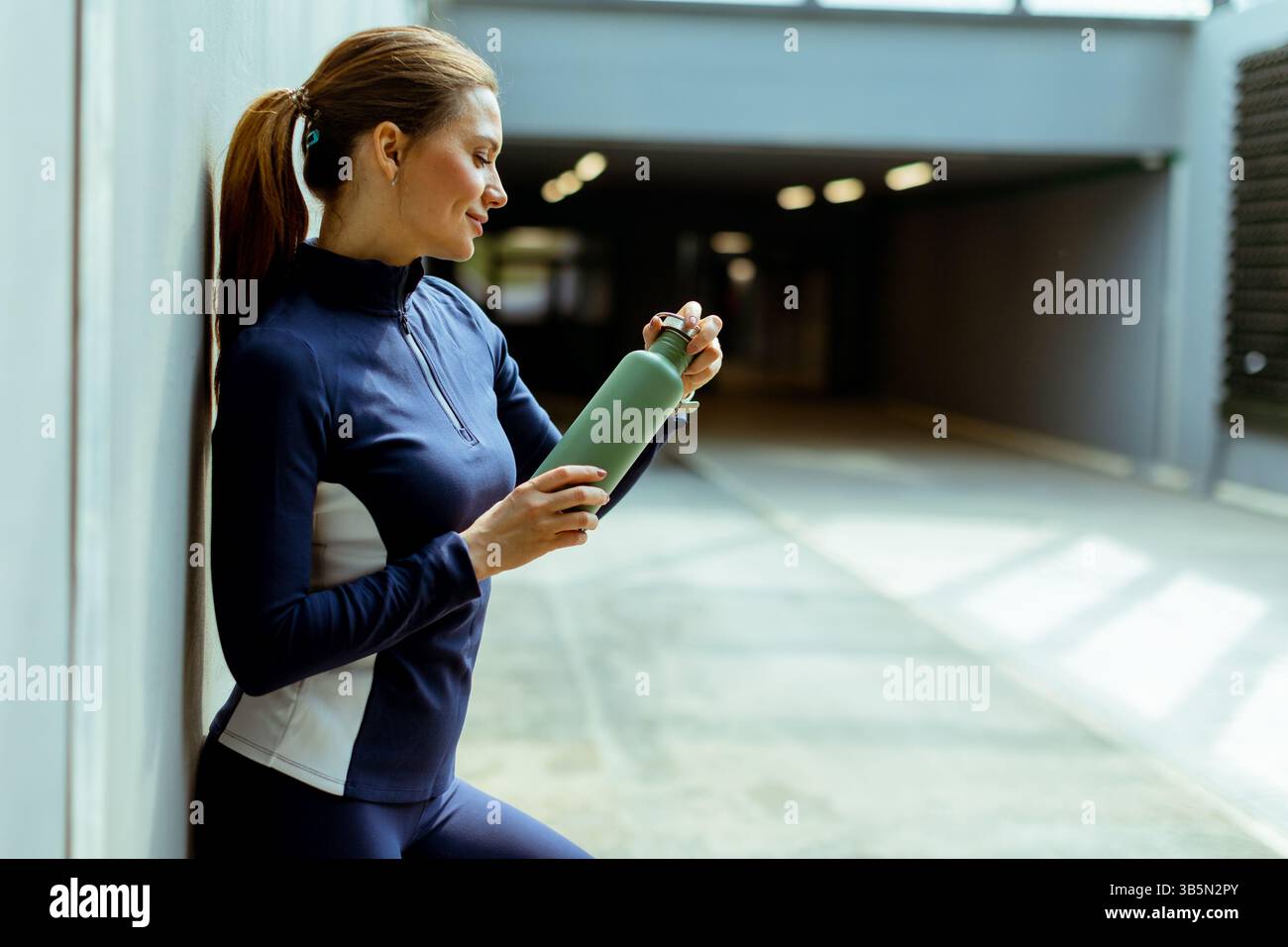 Relaxante dans un espace urbain épuré, une femme en tenue de sport fait une pause pour prendre une gorgée de sa bouteille d’hydratation, appréciant le moment. Banque D'Images