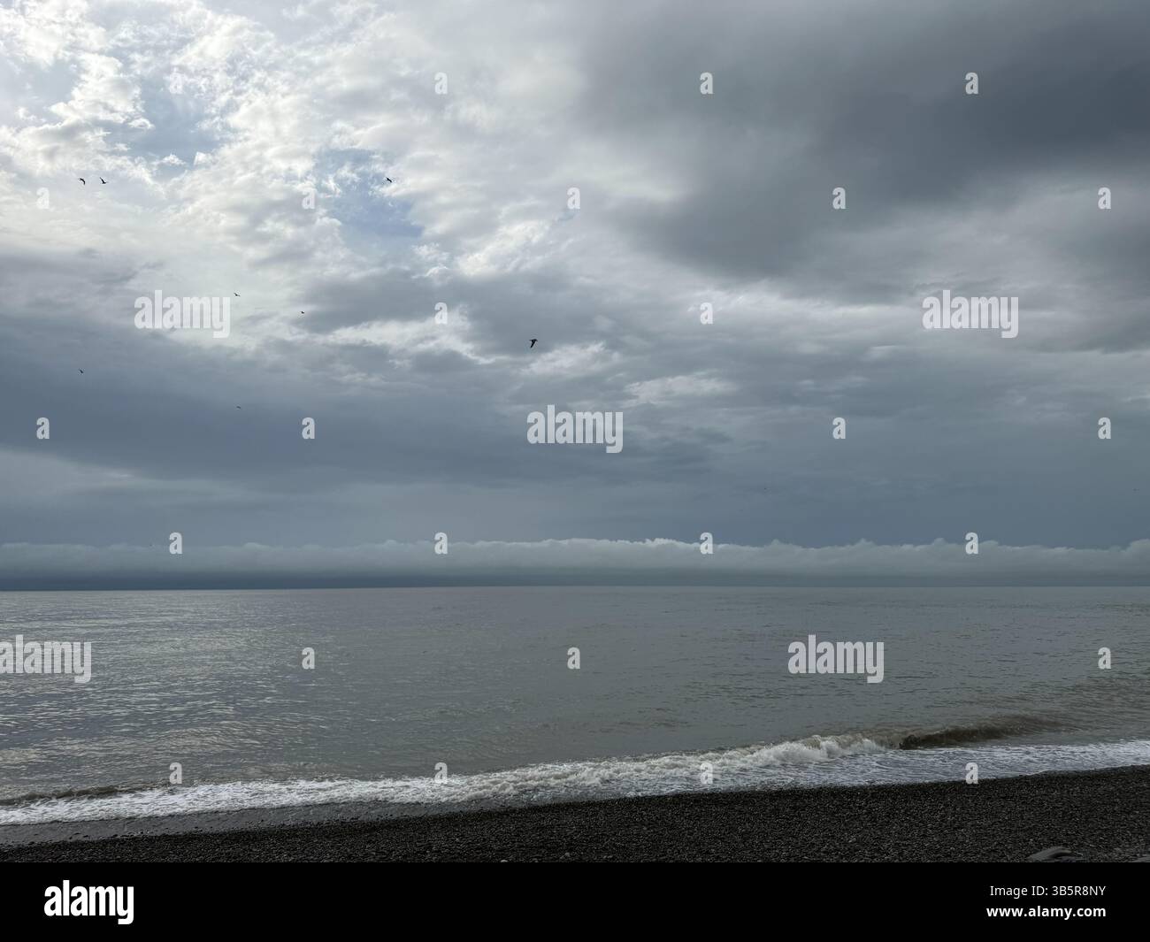 Cette photo capture un paysage marin couvert avec une large étendue de nuages gris au-dessus d'une mer calme et plate. La plage est composée de galets, et de petites vagues g Banque D'Images