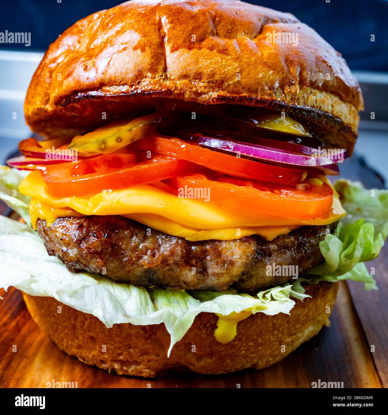 Un hamburger gastronomique est présenté sur une planche de bois, présentant une galette juteuse garnie de fromage fondu, de laitue fraîche, de tomate, d'oignon rouge et de cornichons. Th Banque D'Images