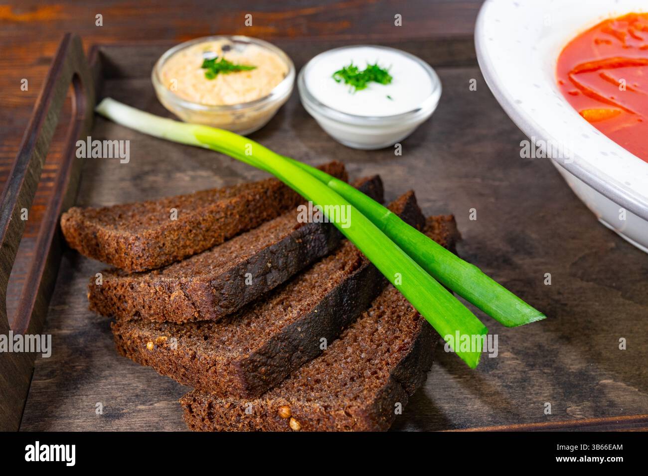 Du pain de seigle noir fraîchement tranché est présenté sur un plateau en bois avec deux trempettes, une crémeuse et une salée, complétées par des tiges d'oignon vert vif, Banque D'Images