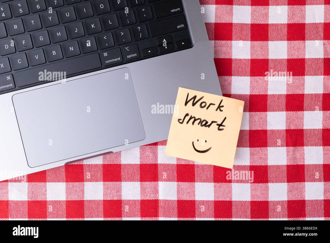 Note autocollante avec message « Work Smart » et smiley placé à côté d'un ordinateur portable sur une surface à carreaux rouge et blanc, symbolisant une motivation simple pour Banque D'Images
