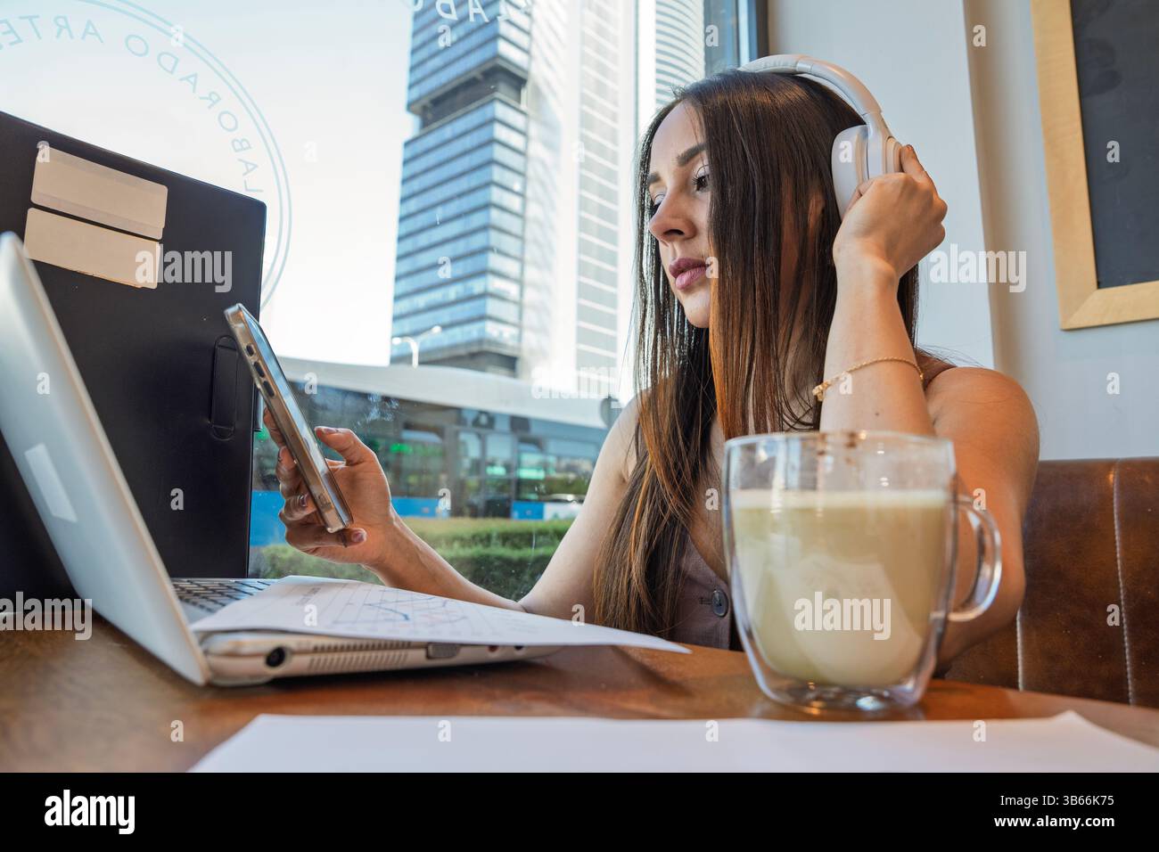 Jeune femme travaillant à distance dans un café, utilisant un ordinateur portable et un smartphone tout en portant des écouteurs et en dégustant une boisson Banque D'Images