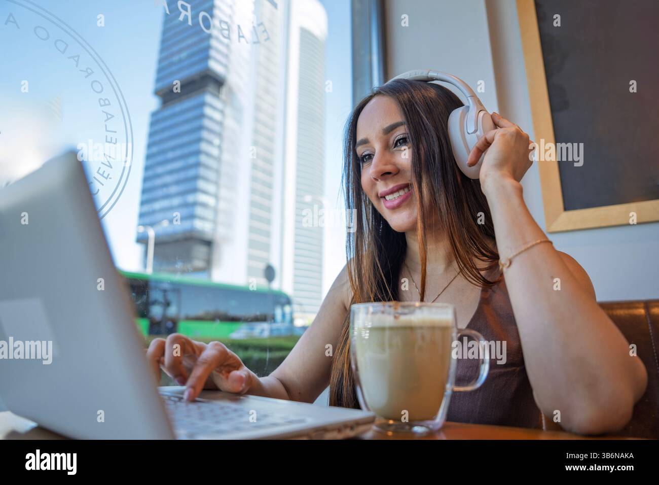 Jeune femme travaillant sur ordinateur portable tout en profitant d'un latte et en écoutant de la musique dans des écouteurs dans un café moderne Banque D'Images