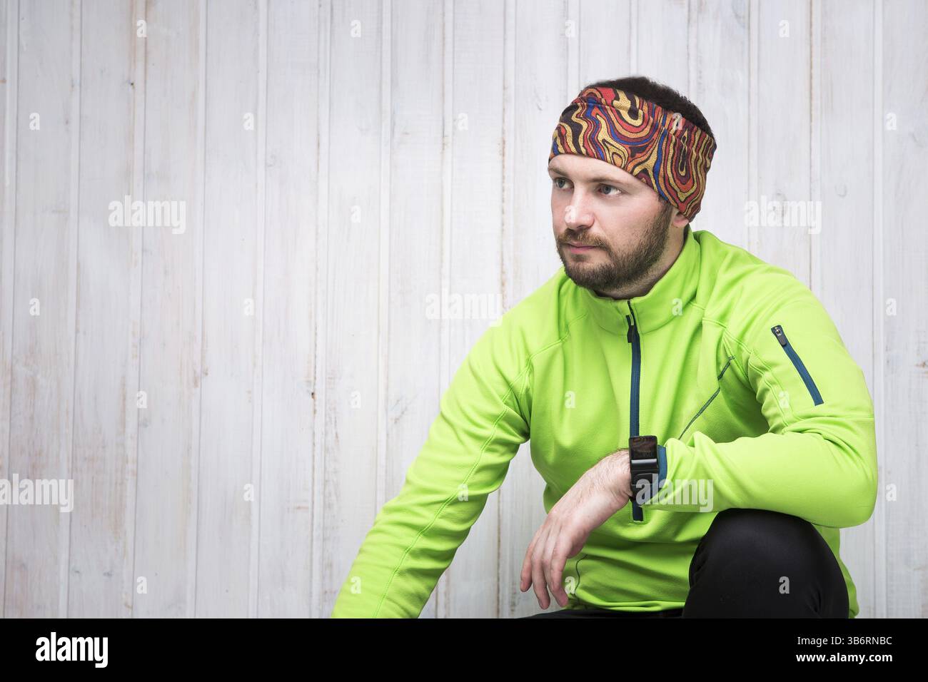 Jeune athlète d'apparence européenne dans une veste verte, montre, pantalon noir, s'assoit. Fond en bois Banque D'Images