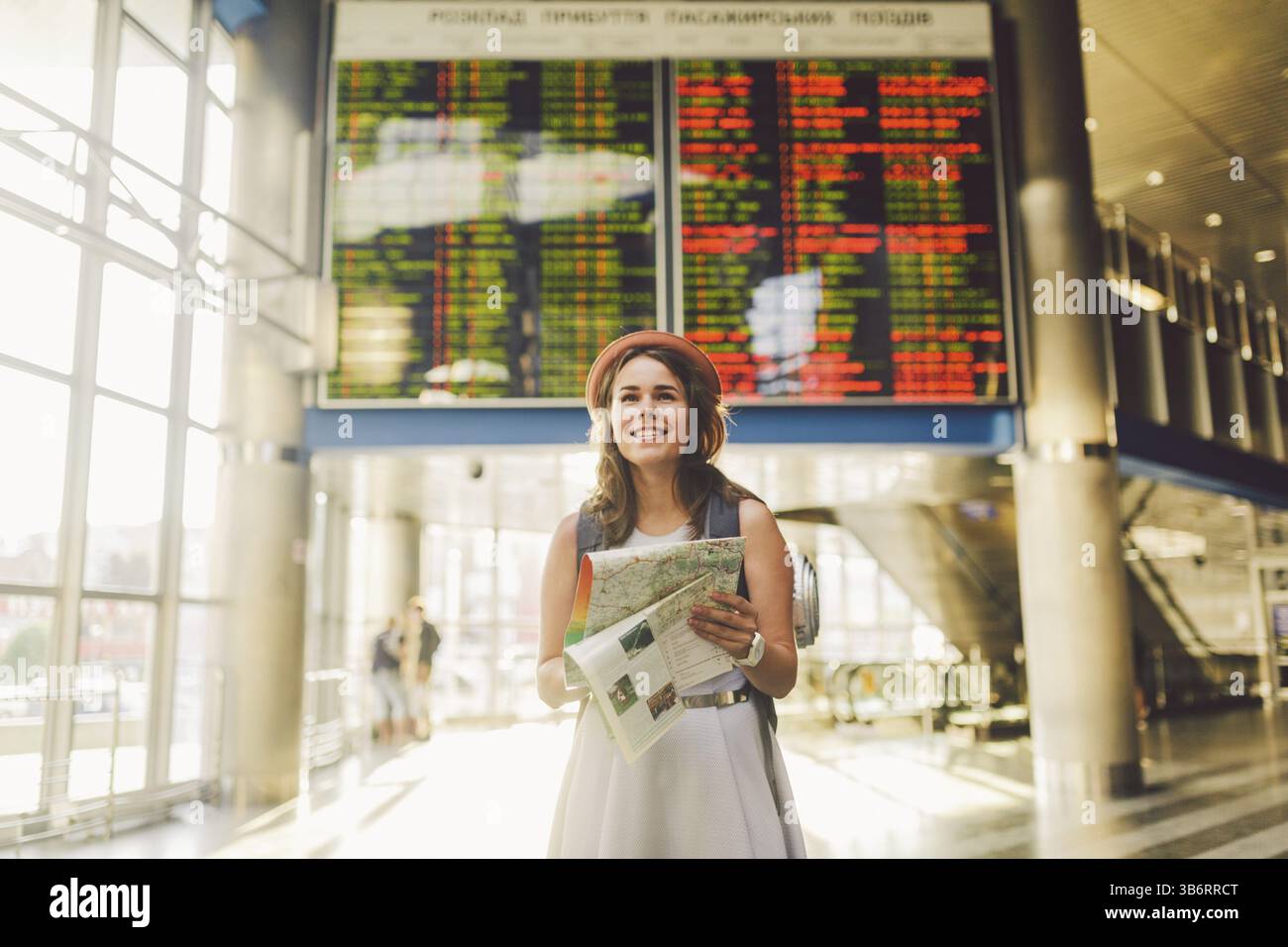 Voyages et transports thématiques. Belle jeune femme caucasienne en robe et sac à dos debout à l'intérieur du terminal de la gare regardant SC électronique Banque D'Images
