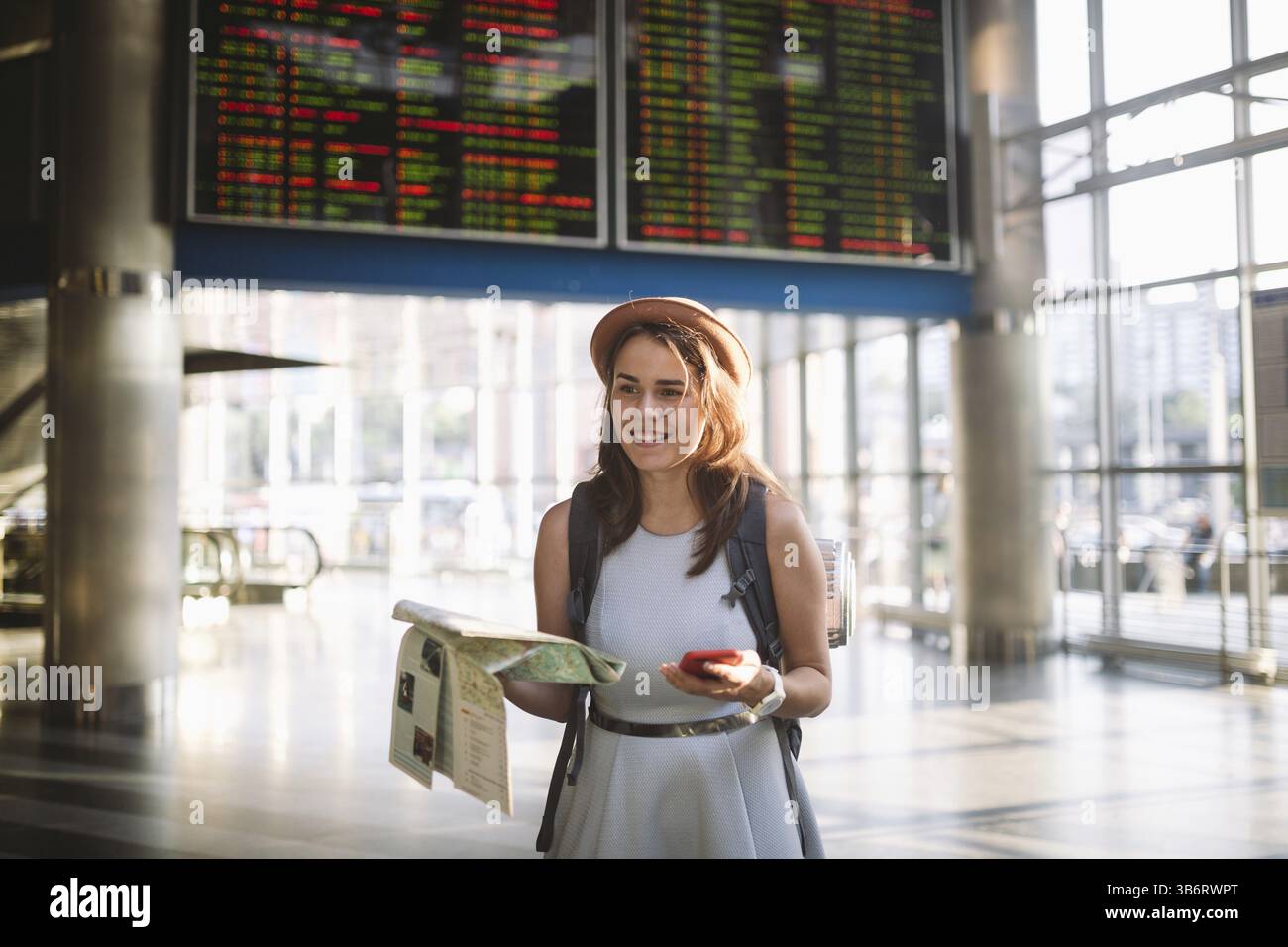 Voyages et transports thématiques. Belle jeune femme caucasienne en robe et sac à dos debout à l'intérieur du terminal de la gare regardant SC électronique Banque D'Images