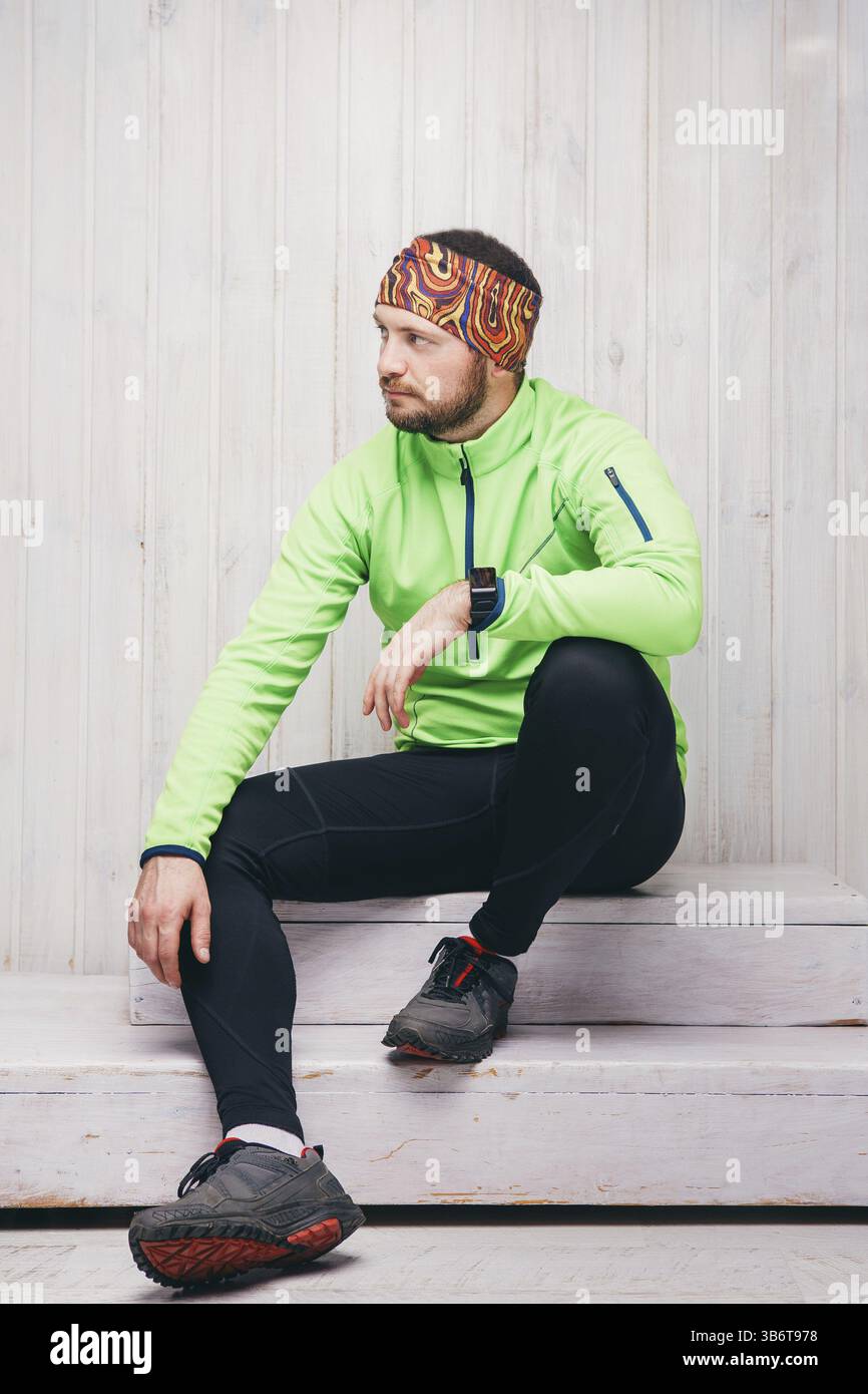 Jeune athlète d'apparence européenne dans une veste verte, montre, pantalon noir, s'assoit. Fond en bois Banque D'Images