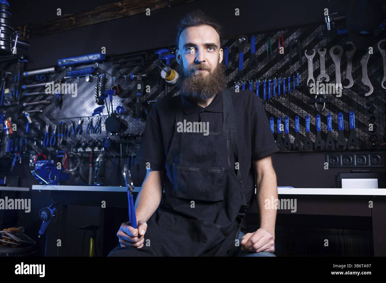 Portrait de propriétaire de petite entreprise de jeune homme à la barbe. Guy atelier mécanicien vélo worker sitting avec l'outil dans sa main dans un vetement noir Banque D'Images