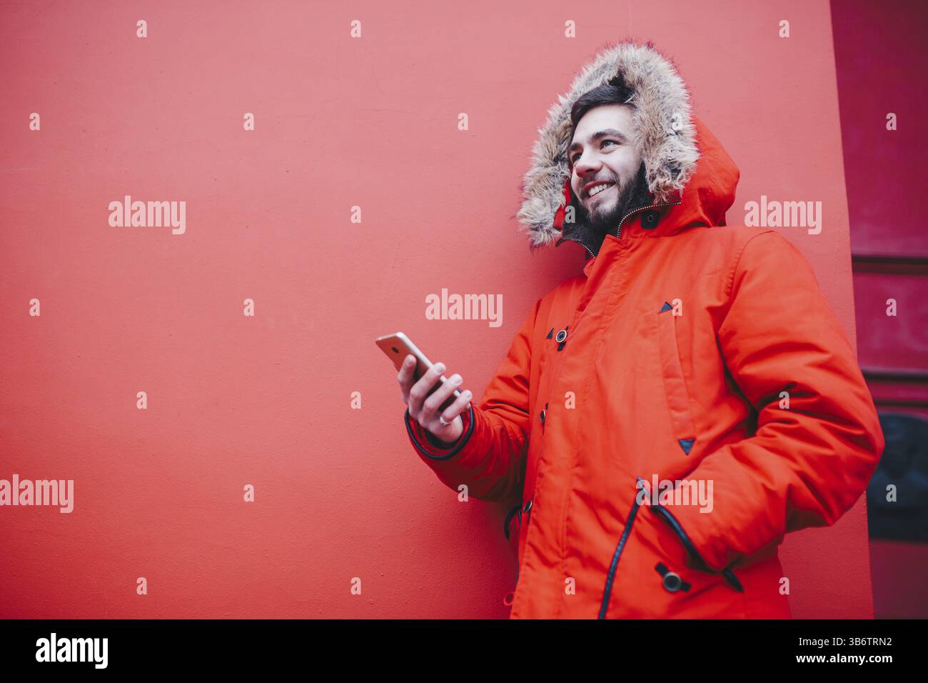 Beau jeune étudiant masculin avec sourire dentelé et barbe se dresse sur fond de mur rouge en veste d'hiver rouge vif avec capuche avec fourrure, utilisations, tient mobi Banque D'Images