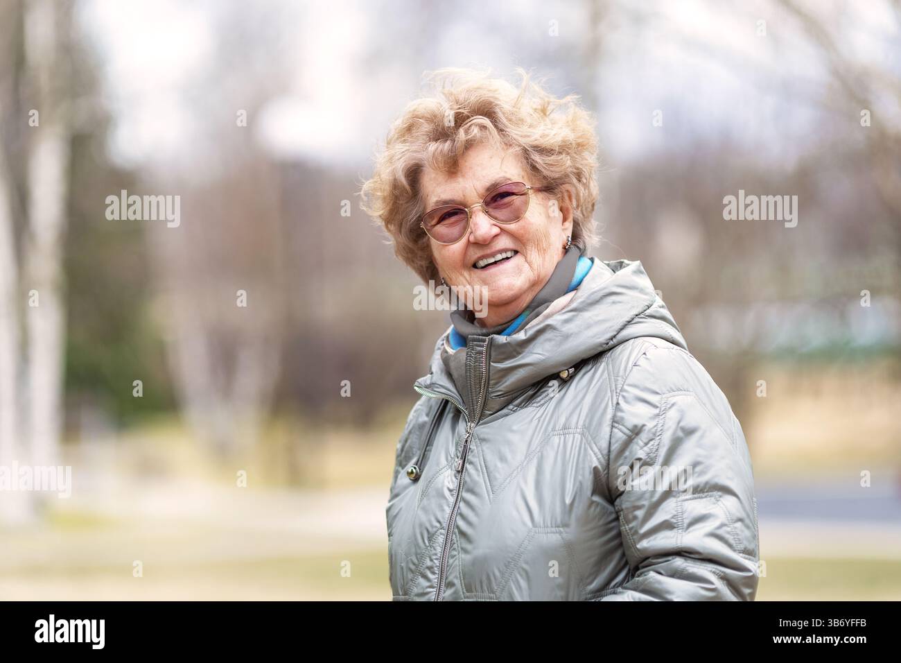 Femme âgée souriante profitant de marcher à l'extérieur au début du printemps, portant une veste chaude et des lunettes, se sentant heureuse, détendue et en bonne santé, montrant une vie active Banque D'Images