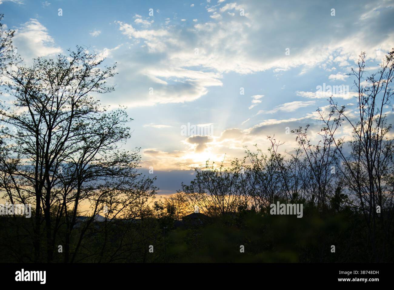 Le soleil se couche derrière des collines avec des arbres. Le ciel est bleu clair, parsemé de nuages. Banque D'Images Le soleil se couche derrière des collines avec des arbres. Le ciel est bleu clair, parsemé de nuages. Banque D'Images