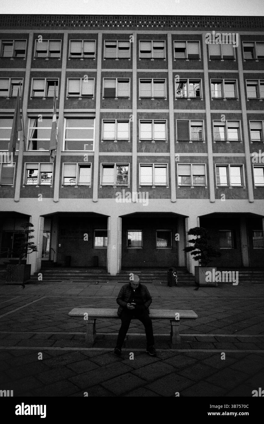 Homme assis sur un banc en béton à l'extérieur de l'architecture de bloc de bureaux des années 60, Italie Banque D'Images