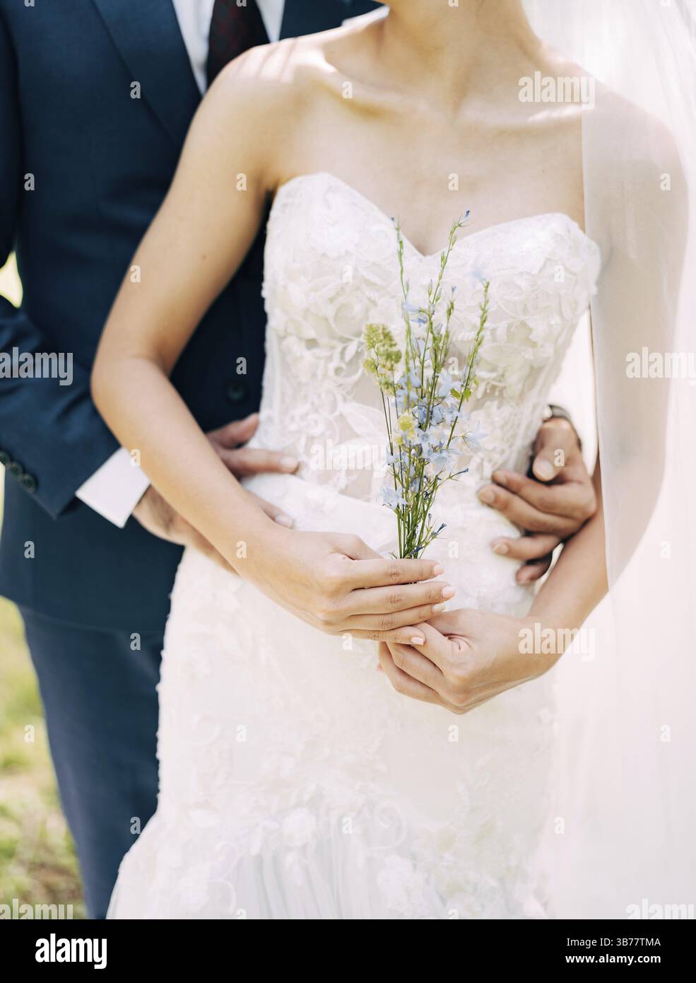 Groom dans un costume bleu embrasse derrière la mariée dans une robe blanche off-the-épaules. Mariée tient des fleurs sauvages dans ses mains. Banque D'Images