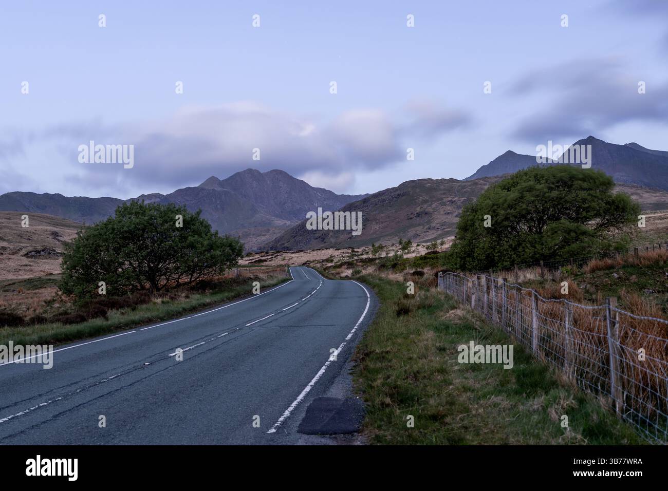 La route sinueuse s'étend vers des montagnes imposantes sous un ciel doux et nuageux, flanqué d'une végétation luxuriante et d'une clôture rustique Banque D'Images